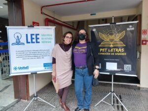 As professoras Flaviane e Kátia Aparecida, posando juntas, cada uma ao lado do respectivo cartaz de seus projetos: LEE e Pet Letras.
