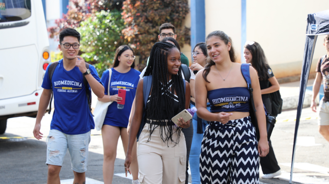 Na imagem, estão, da esquerda para a direita, um estudante oriental, de camisa azul e short jeans; uma estudante preta de camisa azul e calça bege, com cabelos trançados; uma estudante branca de cropped azul com escrito "biomedicina UNIFAL-MG" e uma calça com ziguezague desenhado de preto com fundo branco. Atrás destes estudantes há outros estudantes cuja identificação fica restrita pelo desfoque. Os estudantes estão entrando na Universidade para cursarem o primeiro semestre de 2024, interagindo, sorrindo e em um momento descontraído. Fim da Descrição.