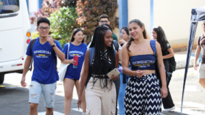 Na imagem, estão, da esquerda para a direita, um estudante oriental, de camisa azul e short jeans; uma estudante preta de camisa azul e calça bege, com cabelos trançados; uma estudante branca de cropped azul com escrito "biomedicina UNIFAL-MG" e uma calça com ziguezague desenhado de preto com fundo branco. Atrás destes estudantes há outros estudantes cuja identificação fica restrita pelo desfoque. Os estudantes estão entrando na Universidade para cursarem o primeiro semestre de 2024, interagindo, sorrindo e em um momento descontraído. Fim da Descrição.